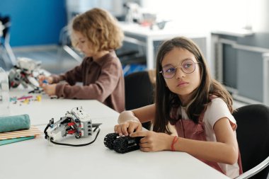 Gözlüklü genç kız derste yeni bir robot modeli geliştiriyor ve masada otururken öğretmenin açıklamasını dinliyor.