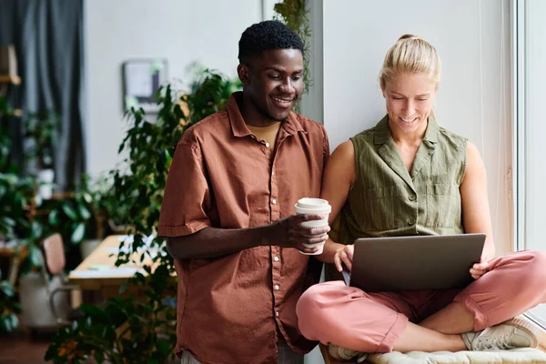 快乐的年轻商人 一边喝咖啡 一边跪在同事的膝上看着屏幕上的笔记本电脑 一边准备演讲 — 图库照片