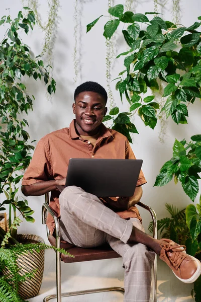 Young smiling employee in casualwear sitting on chair with laptop on his knees and organizing work smong green plants in office