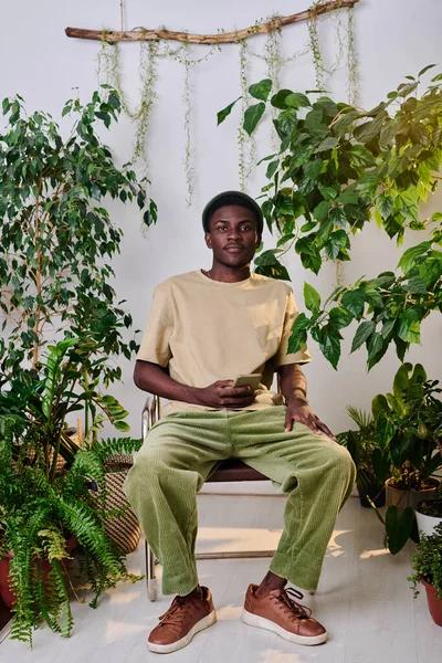 Młody Afroamerykanin Stroju Casualwear Smartfonem Siedzącym Krześle Wśród Zielonych Roślin — Zdjęcie stockowe