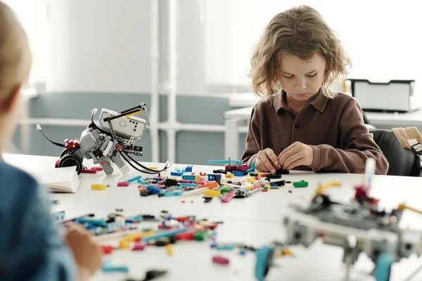 机に座ってロボットの細部をつなぎながら ロボット工学の授業で新しい電子玩具を作る賢い少年 — ストック写真