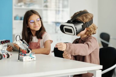 VR kulaklıklı sevimli okul çocuğu, elinde tabletle sınıf arkadaşının yanında oturmuş robot bilimi dersinde ayarlama yaparken sanal oyun oynuyor.