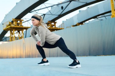 Aktif kıyafetli sarışın genç bir kadın modern mimariye karşı asfalt üzerinde dururken bacak esnetme egzersizi yapıyor.