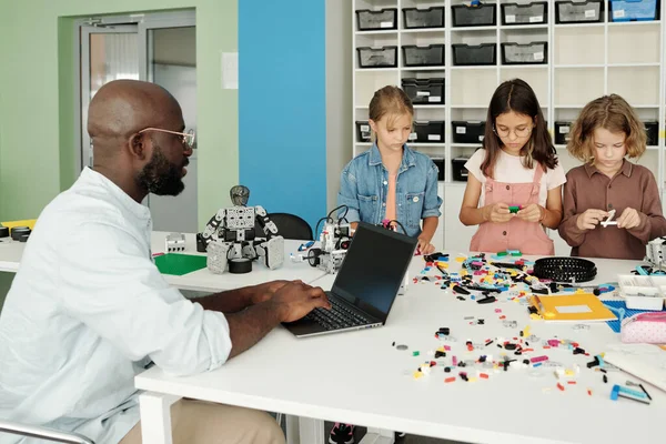 建築家の一部とロボットの作成を接続する小学生のグループの前にテーブルのそばに座っているラップトップを持つロボットの若い教師 — ストック写真