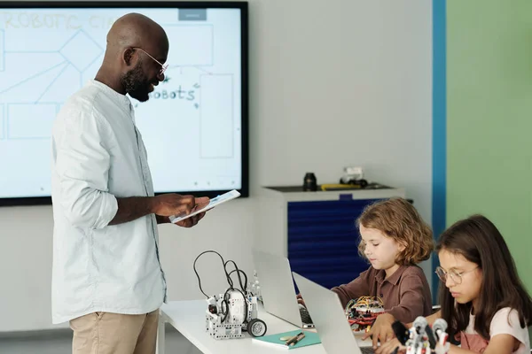 Tabletli Genç Öğretmen Elinde Dizüstü Bilgisayarlarla Okul Çocuklarının Önünde Duruyor — Stok fotoğraf