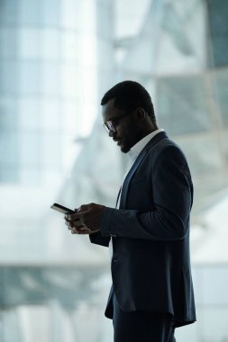 Zarif takım elbiseli modern iş adamının kameranın önünde pencereye karşı dururken akıllı telefonuyla mesajlaşmasının yan görüntüsü.