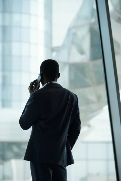 大きなオフィスの窓の前に立って誰かと通信することによって耳でスマートフォンで若い白い襟の労働者のバックビュー — ストック写真