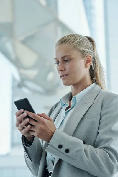 自信的金发女商人站在办公室摄像机前看着智能手机屏幕进行在线交流 — 图库照片