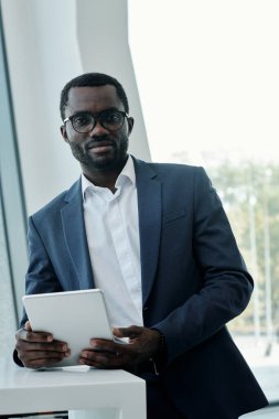 İş yerinde durup tablet kullanırken kameraya bakan takım elbiseli ve gözlüklü Afro-Amerikan genç broker.