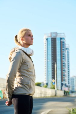 Gri spor ceketli, aktif genç bir kadın modern binaya karşı kentsel çevrede kameranın önünde duruyor.