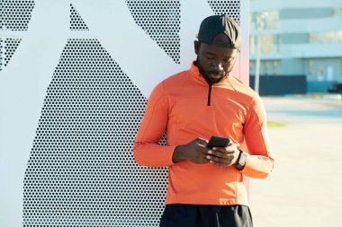 Modern bina duvarının yanında duran akıllı telefonlu genç sporcu mesajlaşıyor ya da kişi listesine bakıyor