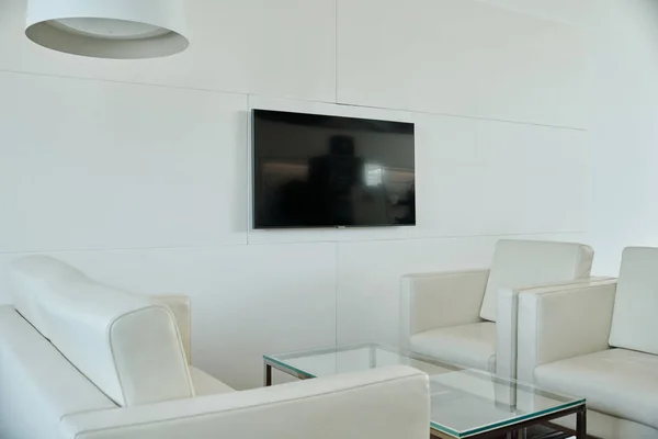White leather couch and two armchairs surrounding rectangular glass table standing by wall with black tv screen in openspace office