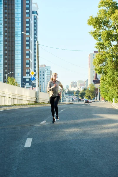 身穿运动服的年轻女运动员在柏油路上奔跑 面对着现代建筑和蓝天 — 图库照片