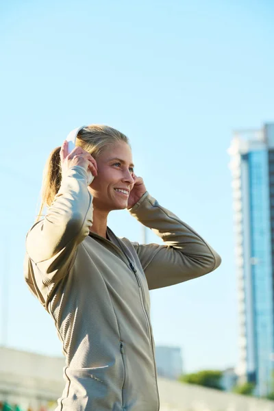 街のカメラの前に立っている間 ヘッドフォンで音楽を聴いている若い笑顔のスポーツ女性の側面図 — ストック写真