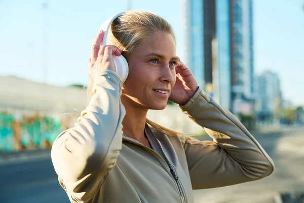 幸せな若いブロンドのスポーツ女性のサイドビュー彼女の頭の上にヘッドフォンを触れながら 都市部の環境で音楽を聴く — ストック写真