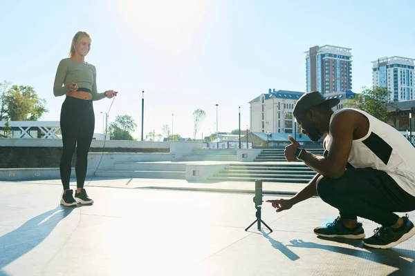若いアフリカ系アメリカ人のスポーツマンは ブロンドの女性選手のスポーツトレーニングのビデオを撮影する前にスマートフォンの画面を指して — ストック写真