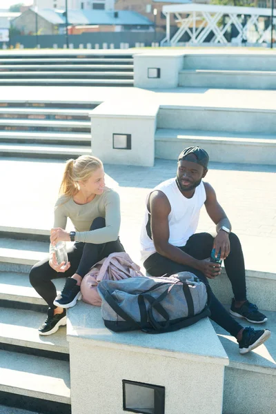スポーツウェアの若い異人種間の選手は トレーニングやジョギングの後 屋外で階段に座って水を持っているとチャット — ストック写真