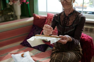 Koltuğunda boş boş otururken ve çapraz dikiş atarken beyaz bez ile ince ahşap halkalar tutan yaşlı kadının elleri.