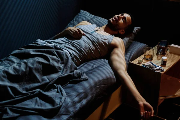 年轻而有压力的男人躺在床头柜边试图入睡 床头柜里装着威士忌 安眠药和照片 — 图库照片