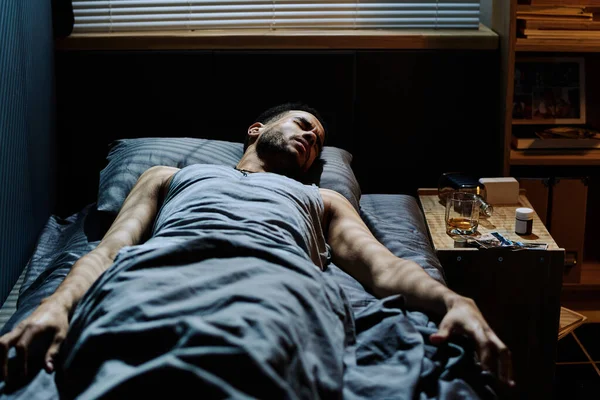 一个患有创伤后综合症的疲倦的年轻人躺在床头柜旁 一边喝着威士忌和药瓶 一边睡觉 — 图库照片