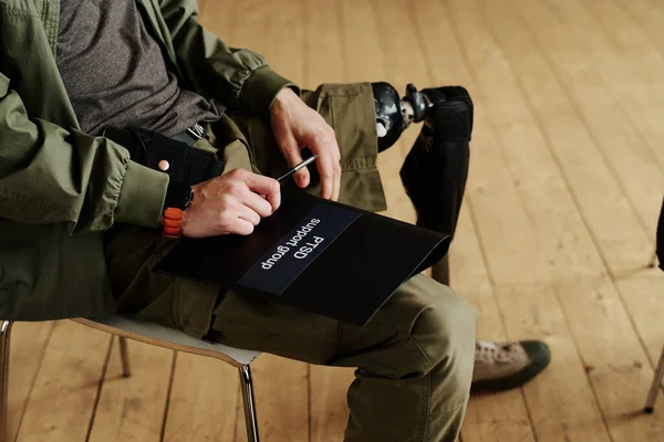 stock image Young man with disability holding black folder of PTSD support group while sitting on chair during psychological session