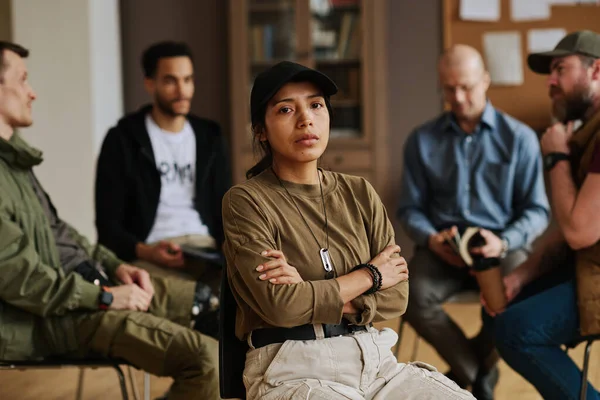 Young stressed Hispanic woman with her arms crossed on chest looking at camera while sitting against intercultural men having discussion