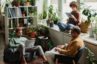 Gündelik kıyafet giyen üç çok kültürlü iş arkadaşı ofiste mobil cihazlar kullanırken olgun bilişim mühendisleri bilgisayar ekranında veri çözüyor.
