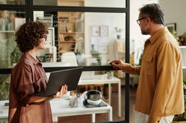 Kendine güvenen olgun bir adam notlarla şeffaf tahtayı işaret ederken onları dizüstü bilgisayarlı genç bayan meslektaşına açıklıyor.