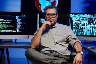 Akıllı günlük giyimli ve gözlüklü, sunucu odasında bilgisayar monitörlerine karşı oturan ciddi, olgun bir erkek yazılım geliştiricisi.