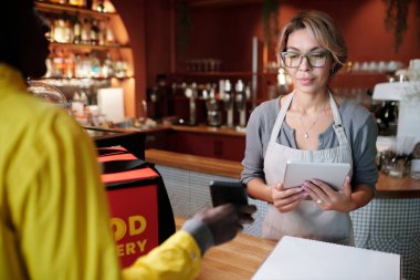 Çevrimiçi menü ve çevrimiçi sipariş listesini kontrol ederken kurye tarafından tutulan akıllı telefon ekranına bakan tabletli genç garsona odaklan