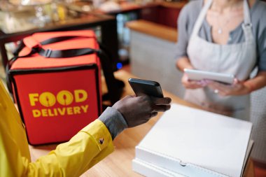 Büyük kırmızı çantalı akıllı telefonu elinde tutan kuryenin eli pizza dolu kutular ve garson kıza online menü öğelerini gösteriyor.