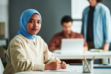 Mavi tesettürlü genç Müslüman kadın kameraya bakarken ya da gramer testini iş yerinde iki erkeğe karşı yaparken bakıyor.