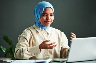 Gündelik kıyafet ve mavi tesettürlü kendine güvenen genç Müslüman kadın çevrimiçi ders sırasında dizüstü bilgisayarın önünde otururken bir şey anlatıyor.