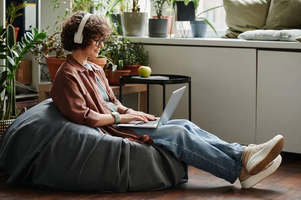 穿着耳机和便服的年轻女性员工坐在现代办公室的软垫椅上 一边听音乐 一边上网 — 图库照片