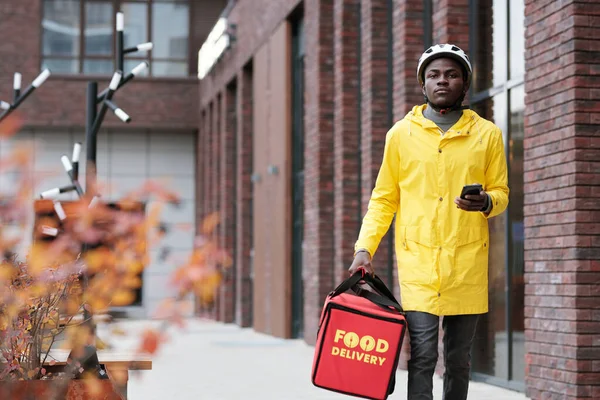 建物のレンガの壁に沿って歩いている間に大きな赤い袋を運ぶヘルメットと黄色のジャケットでの食品サービスの若いアフリカ系アメリカ人の宅配業者 — ストック写真