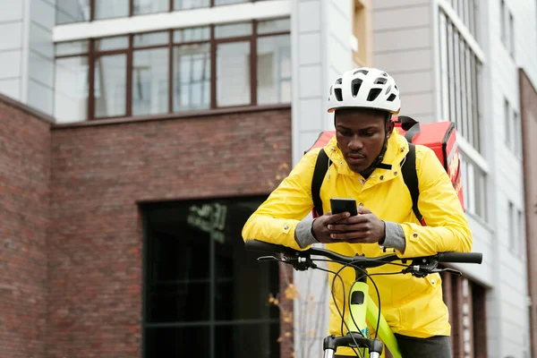Junger Afroamerikanischer Auslieferer Mit Handy Sitzt Auf Dem Fahrrad Und — Stockfoto