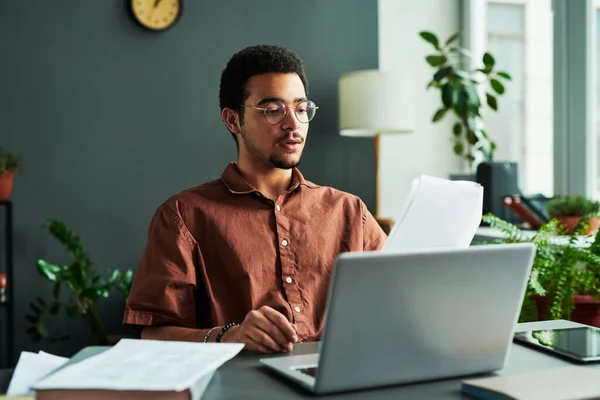 Young confident man with papers sitting by workplace in front of laptop and making report or explaining new information to online audience