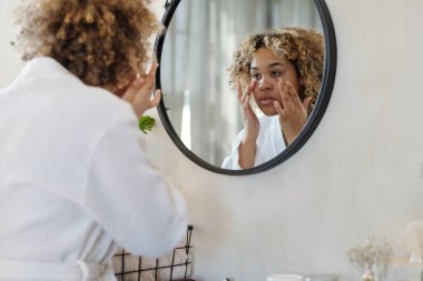 Duvarda aynanın önünde duran ve sabah duştan sonra göz altına nemlendirici krem süren genç bir kadın.