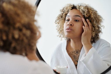 Genç Afrikalı Amerikalı bir kadın, cildine losyon sürerken kendini arındırma ürünüyle yüzünü nemlendiriyor.