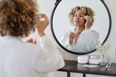 Bornozlu genç bir kadın, yuvarlak aynanın önünde dikilirken temizleyiciyle temizledikten sonra yüzüne bir damla gençleştirici serum sürüyor.