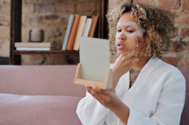 Bornozlu genç kadın yatakta oturup aynaya bakarken cilt bakımı sırasında yüzüne kuvars silindiriyle masaj yapıyor.