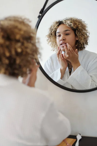 若いアフリカ系アメリカ人の女性は鏡を見ながら朝のルーチン中に彼女の顔に保湿クリームを適用します — ストック写真