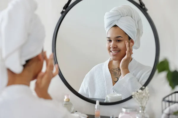 朝のシャワーの後 浴室の鏡を見ながら顔に保湿クリームを適用頭の上にタオルで幸せな若い女性 — ストック写真