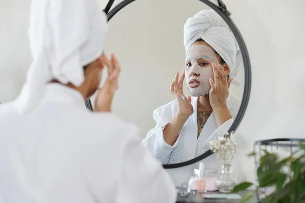 stock image Young woman in white bathrobe putting hydrating sheet mask on her face while standing in front of mirror after having morning shower