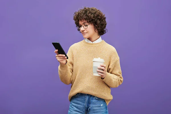 年轻女子 一头乌黑的短卷发 独立地透过手机照片 在紫色背景下喝咖啡 — 图库照片