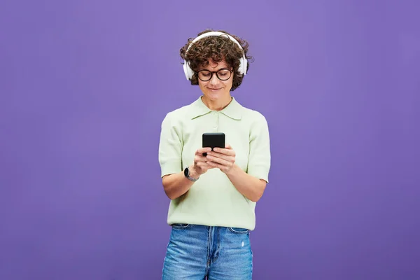 Young woman in casualwear and headphones scrolling in mobile phone while choosing new sound track in playlist against violet background