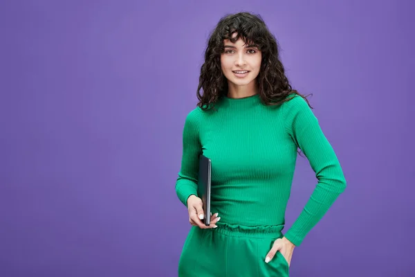 若いです自信のある女性従業員または学生で緑のカジュアルウェア保持ラップトップとカメラを見ながら紫の背景に立って — ストック写真