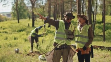 Orta uzunluktaki Asyalı ve beyaz kadın çevreciler üniformalarını giyip tablet bilgisayar kullanıyorlar, konuşuyorlar, güneşli bahar günlerinde ormanda duruyorlar.