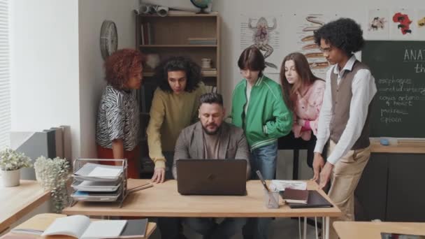 五花八门的高中学生围绕着坐在课桌前的男教师站着 下午使用笔记本电脑 — 图库视频影像