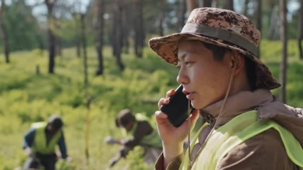 在阳光明媚的夏日 年轻的亚洲女林务员站在森林里 一边用手机聊天 一边进行胸部检查 — 图库视频影像
